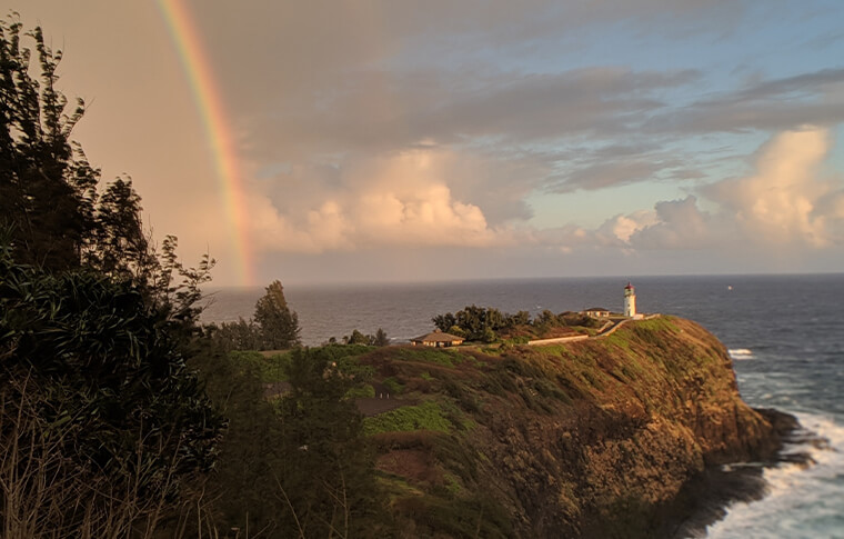 Sunset and rainbow with a lighthouse perched at the end of the cliff