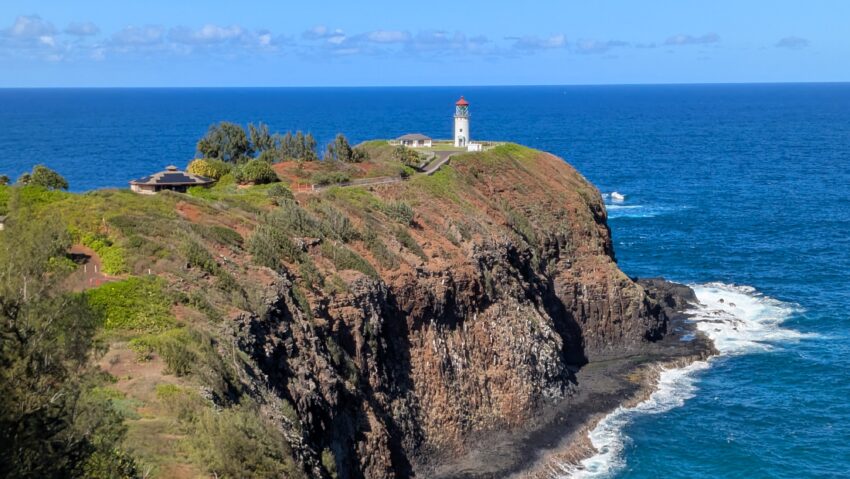 A lighthouse stands on a green cliff in Kauai, overlooking a vast blue ocean under a clear sky. With sunny weather and waves crashing against the rocky shoreline, it's the perfect getaway.