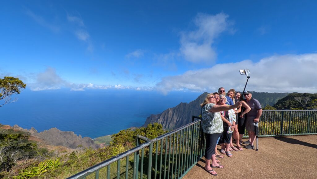 A group of six people takes a selfie with a stick at a breathtaking Kauai viewpoint, overlooking mountains and the ocean under the clear blue sky—capturing the essence of this perfect getaway in sunny weather.