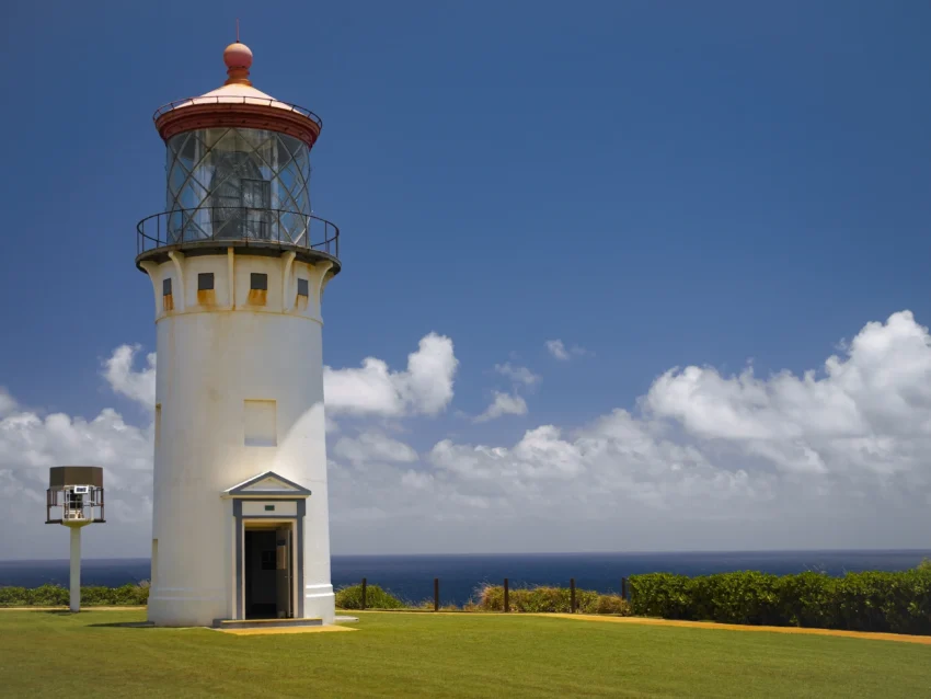 The Kīlauea Lighthouse, with its white tower and red top, stands on a grassy lawn near a small building—one of Kauai’s beloved local spots, where people stroll beneath a partly cloudy sky.