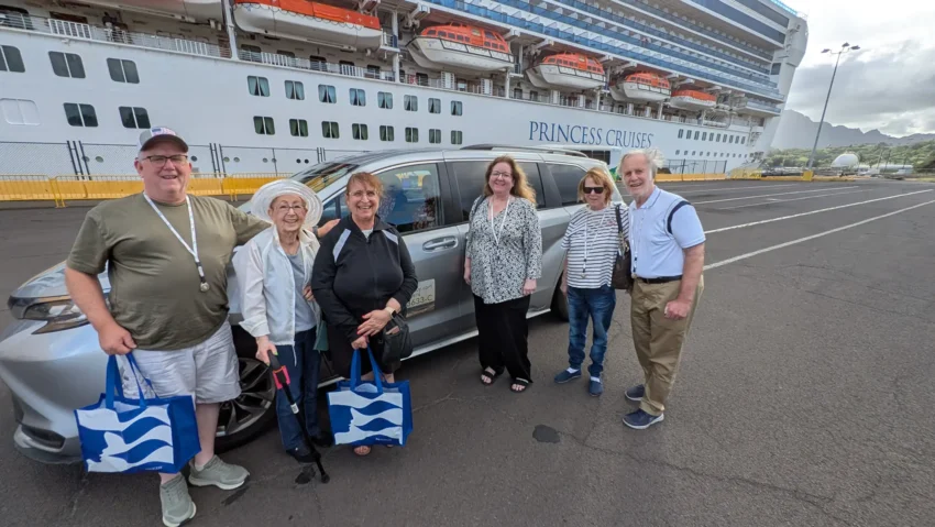Six people stand in front of a minivan in a parking lot with a Princess Cruises ship docked in the background. Some hold blue and white tote bags, ready for their Kauai road trip and eager to share Kauai travel tips.