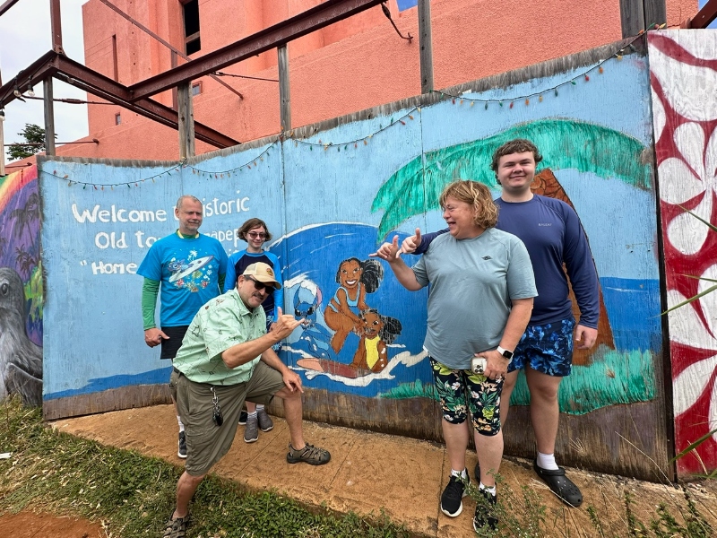Doug posing with the shaka next to a group of guests standing by a colorful tropical mural